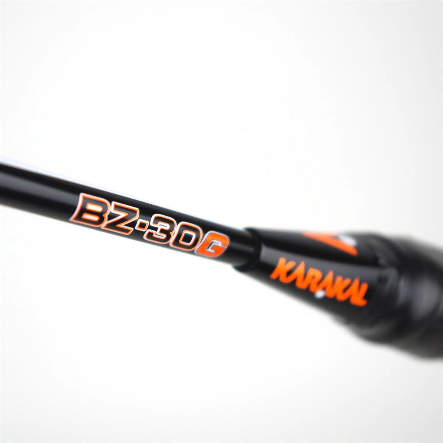 Close-up of a black handle from the Karakal BZ 30 2.1 Badminton Racket, featuring orange and white text that reads "BZ-30G" and "KARAKAL." The handle, part of the Karakal series, is slightly angled against a faded white background to emphasize the branding.