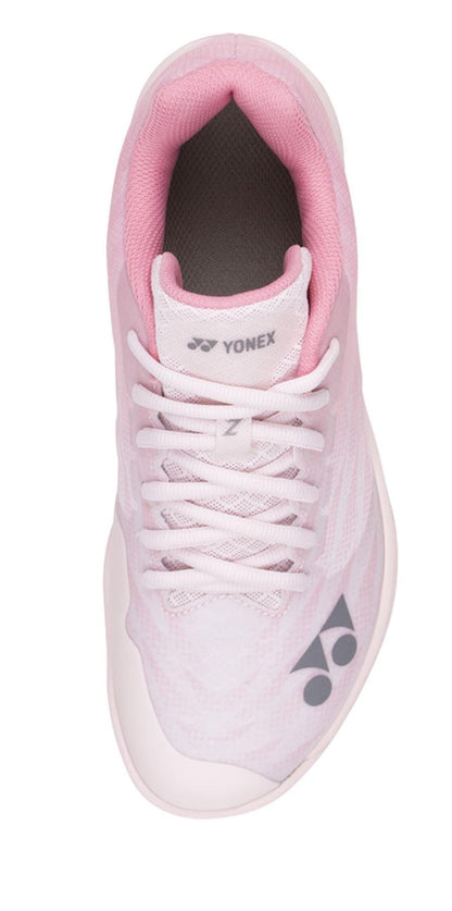 A top-down view of the Yonex Power Cushion Aerus Z2 Women's Badminton Shoe in light pink with white laces showcases the Yonex logo on the tongue and side. The shoe features a rounded white sole utilizing Power Cushion technology and a subtle mesh texture on the upper fabric, complemented by Durable Skin Light for enhanced longevity.