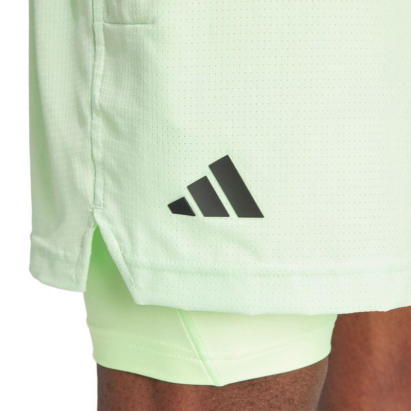 Close-up of the light green ADIDAS Melbourne Mens 2in1 Badminton Shorts - Green by adidas, featuring a black logo on the side. The fabric looks breathable and slightly textured due to HEAT.RDY technology. Underneath, you can see matching light green leggings crafted from recycled materials.