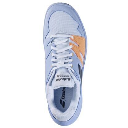 Aerial perspective of a Babolat Shadow Team 2 women's badminton shoe in light blue and orange, complete with white laces. The shoe displays the Babolat brand name on the tongue and side, featuring a mesh-textured upper and a durable Michelin rubber outsole for superior grip.