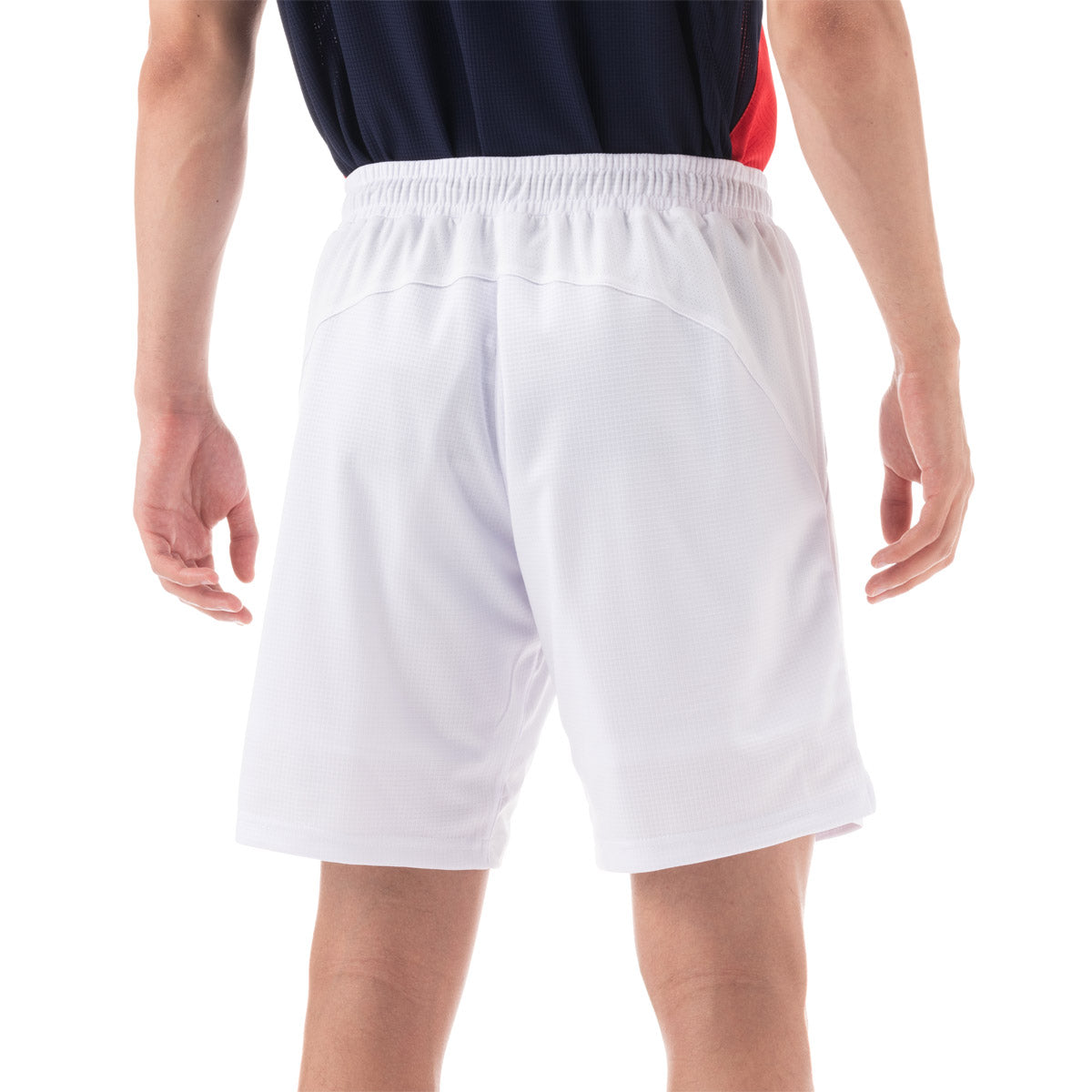 A person stands with relaxed hands, wearing a dark sports jersey paired with the Yonex 15131 Men's Shorts in white. The white background accentuates the shorts' UV reduction and VERY COOL technology, guaranteeing comfort and style in any setting.