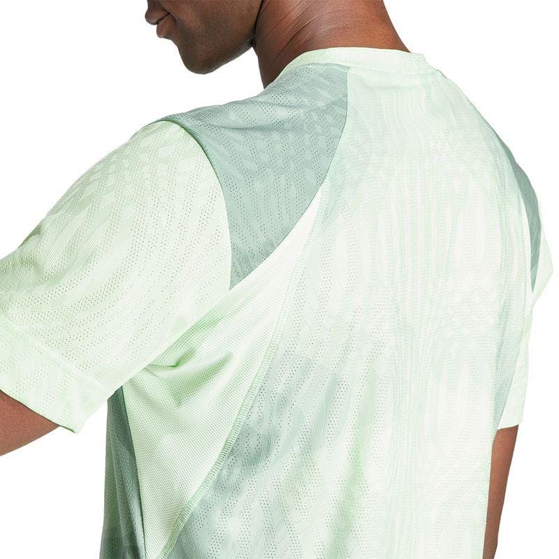 The individual is dressed in a light green ADIDAS Melbourne Men's Pro Badminton T-Shirt, crafted from recycled polyester and featuring a subtle pattern. The image highlights the upper back and shoulders, emphasizing the shirt's design as the person turns slightly to the side to showcase this adidas creation beautifully.