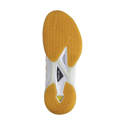 The image displays the sole of a Yonex Power Cushion 65Z3 Women's Badminton Shoe in a mustard yellow color with white sections. It features a Yonex Power Cushion design, complete with grooves and textured surfaces for traction, as well as a small triangular detail in the center, ensuring an ultra-comfortable fit.