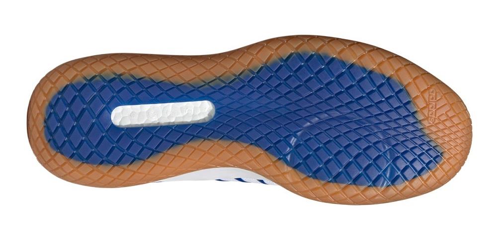 The image displays the sole of an ADIDAS Stabil Next Gen Primeblue men's badminton shoe, ideal for indoor court activities. It showcases a blue tread with a diamond pattern and includes a BOOST-enhanced midsole section, while the outer edge features a brown translucent border.