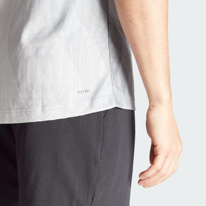 A person wearing an ADIDAS Melbourne Men's Pro Badminton T-Shirt in grey is seen from the back. The shirt is crafted with Parley Ocean Plastic and has subtle "HEAT.RDY" text near the hem. The background is a pale gray color.