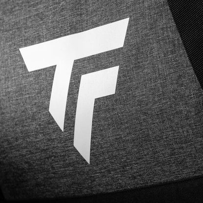 The white, stylized "TF" logo adorns a textured dark gray fabric surface, evoking the look of the Tecnifibre All Vision Badminton Backpack - Grey. The backdrop hints at a racquet sports theme, possibly part of a wide-angle opening on a garment or accessory.