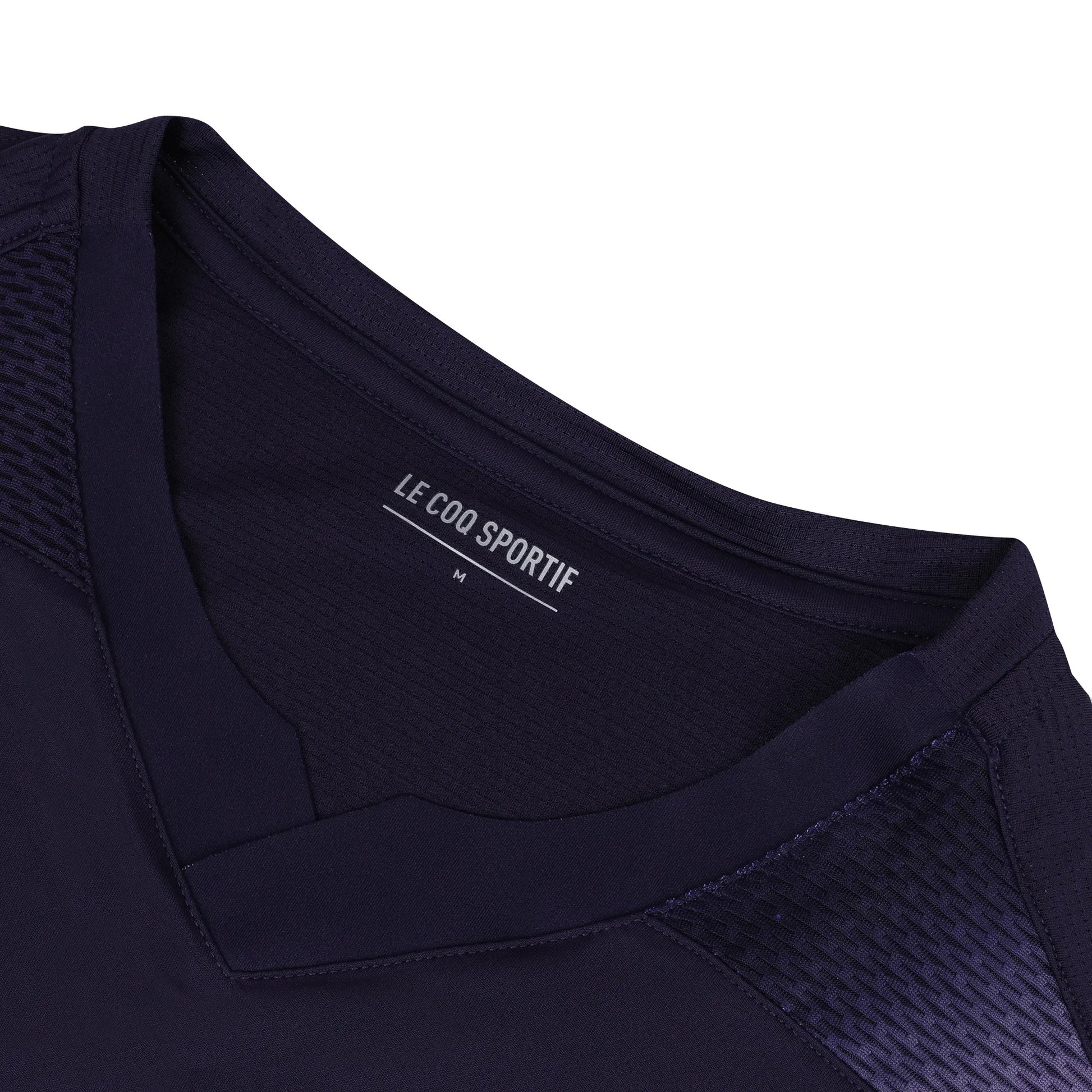 Close-up of a deep purple V-neck badminton t-shirt with the Le Coq Sportif label inside the collar, displaying size "M." The breathable polyester fabric ensures fluidity of movement and features textured detailing on the shoulder.