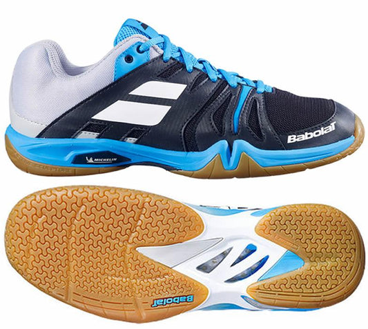 Introducing the Babolat Shadow Team Badminton Shoes in Black Blue: these shoes showcase a distinct blue and white design with a black upper, complemented by blue laces and accents. They feature an orange-brown textured tread pattern made from Michelin rubber for exceptional grip, with the Babolat brand name prominently displayed on the side and sole.