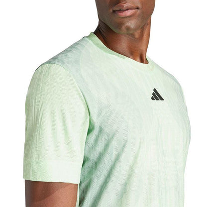 A person is wearing an ADIDAS Melbourne Men's Pro Badminton T-Shirt in green, crafted from recycled polyester with a black logo on the chest, standing against a white background. The view captures only the upper torso and part of the face.