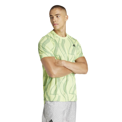 A man stands with arms crossed, wearing an ADIDAS Mens Club Graphic Badminton T-Shirt in green, crafted from recycled materials, paired with light gray shorts. He looks to the side against a plain white background.