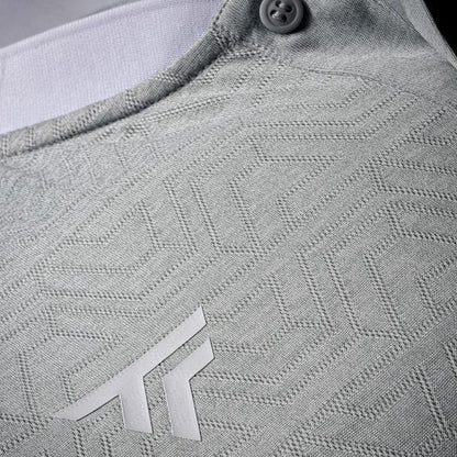 A close-up showcases the textured silver fabric with a geometric pattern, highlighting the small gray logo and button on the collar. Crafted from polyester mesh, the Tecnifibre Men's Team Badminton Mesh Polo Shirt provides optimum breathability for ultimate comfort.