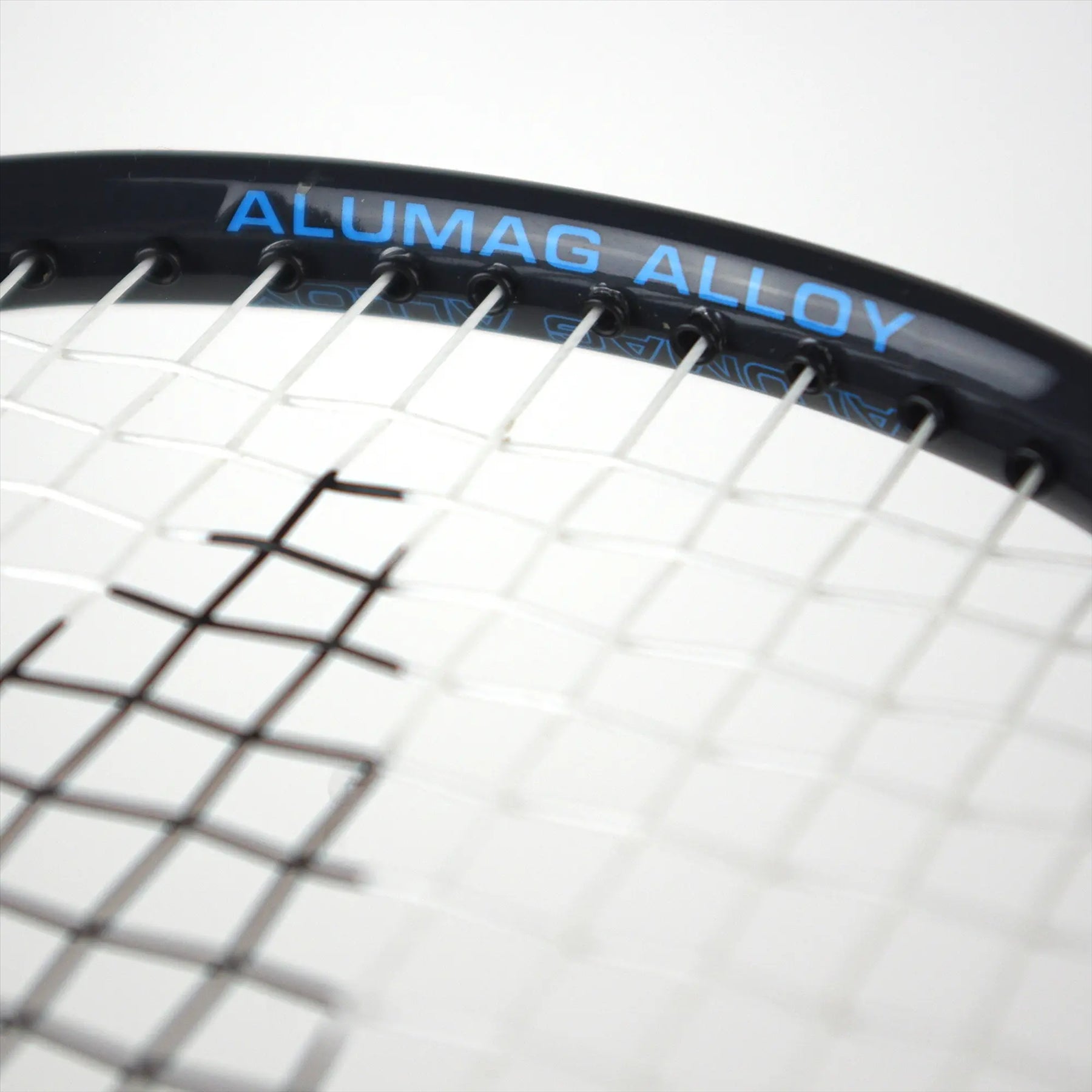 Close-up of a tennis racket with white strings. The frame is black, featuring "ALUMAG ALLOY" in blue. With an isometric head design similar to the Karakal CB-3 2.1 Badminton Racket, the focus remains on the top part of the racket head, making it ideal for beginners honing their skills.