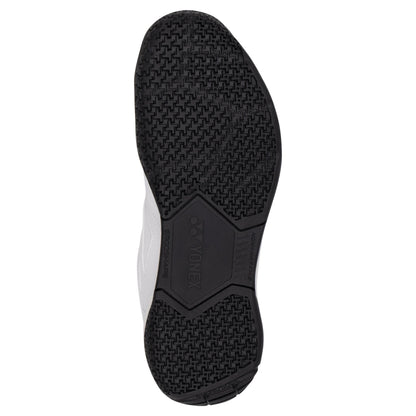 The image shows the sole of a Yonex Power Cushion Strider Ray Men's Badminton Shoe, featuring a black textured rubber pattern for enhanced grip. "YONEX" is prominently displayed at the center. The shoe's upper is white, and the sole showcases various textures and grooved designs, highlighted by Power Cushion technology.