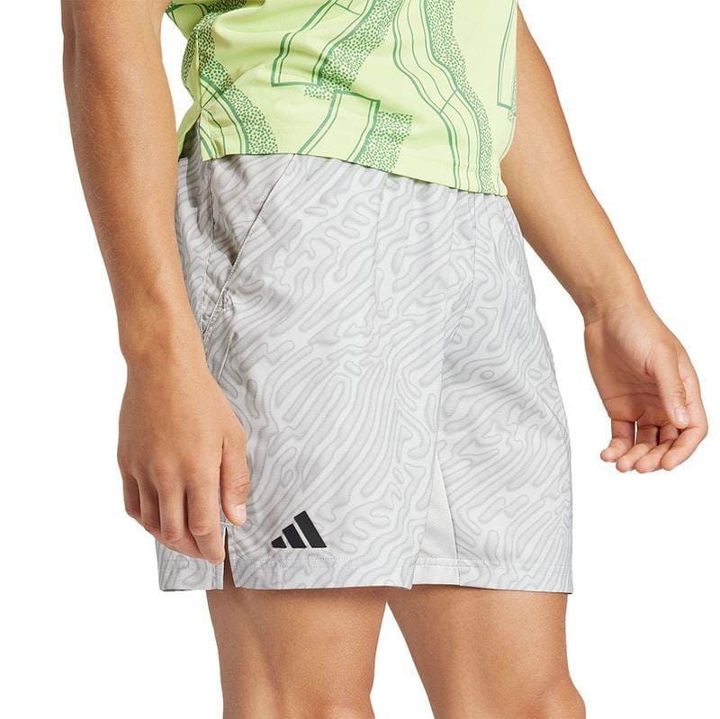 A person is wearing a light green shirt with abstract patterns and light gray ADIDAS Melbourne Men's Pro 7 Inch Badminton Shorts made from recycled polyester, showcasing the HEAT.RDY technology and adorned with a black Adidas logo. Their hands rest casually by their sides.