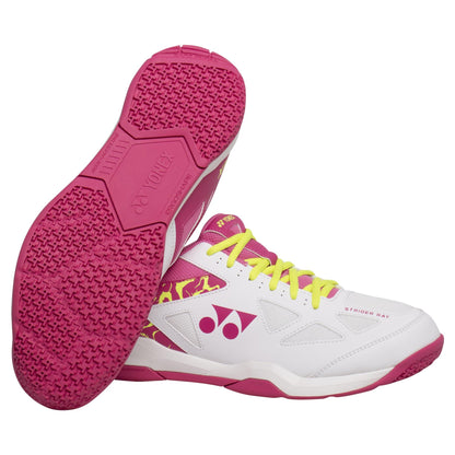 A pair of Yonex Power Cushion Strider Ray Women's Badminton Shoes in a white and pink color scheme, featuring the distinctive Radial Blade Sole technology on the pink soles. They come with yellow laces and display a pink logo along with a decorative design on the sides. One shoe stands upright, while the other displays its pink patterned sole that provides Power Cushion comfort.