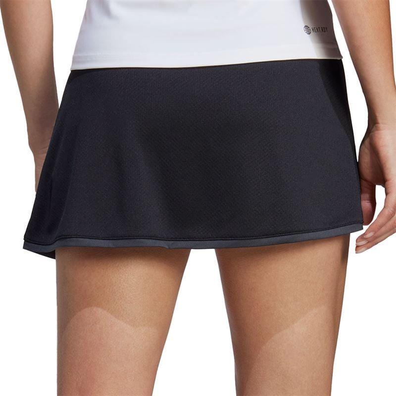 A person wearing a white top and the ADIDAS Women's Club Badminton Skirt - Black with AEROREADY technology and a comfortable wide waistband stands with their back to the camera. The skirt's smooth, slightly flared fabric contrasts elegantly against the plain white background.