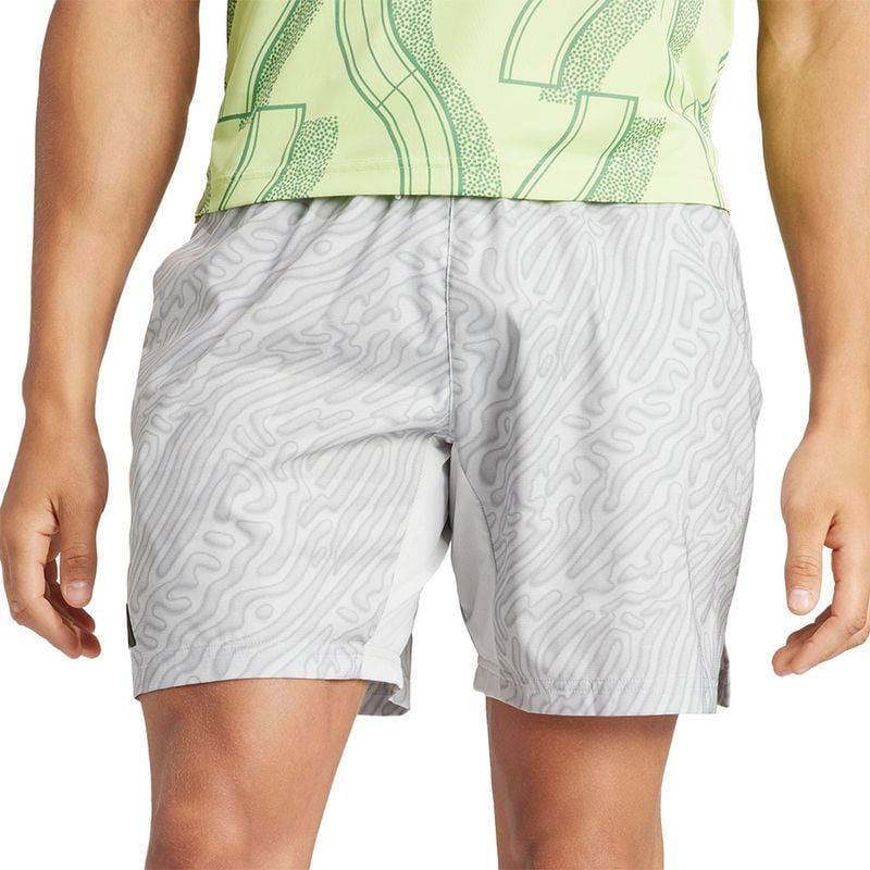 A person is dressed in the adidas Melbourne Men's Pro 7 Inch Badminton Shorts in grey, crafted from recycled polyester and showcasing a wavy line design. They pair these shorts with a light green top embellished with abstract patterns. The focus is on the shorts for their advanced HEAT.RDY technology, set against a plain white background.