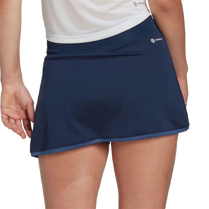 The image depicts a person wearing an adidas Women's Club Badminton Skirt in navy, combined with a white top, viewed from behind. The skirt includes built-in short tights and has a small logo on the side. The background is plain.