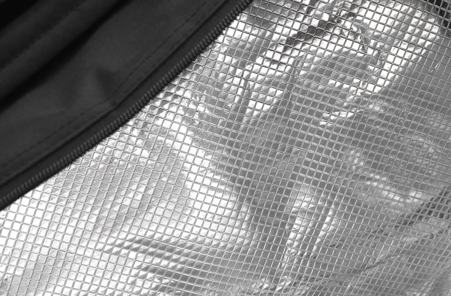 Close-up of a reflective silver foil material with a textured, grid-like pattern, partially framed by the black fabric border of a Dunlop CX Performance 12 Racket Thermo Bag in Black/Red. The surface reflects light, creating contrast between the shiny foil and matte edging, reminiscent of the sleek design characteristic of Dunlop's CX Performance gear.