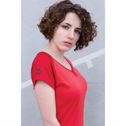 A person with curly brown hair, wearing a red Babolat Play Women's Cap Sleeve Badminton Top in Tomato Red, crafted from breathable fabric, stands against a light gray background, looking at the camera with a neutral expression.