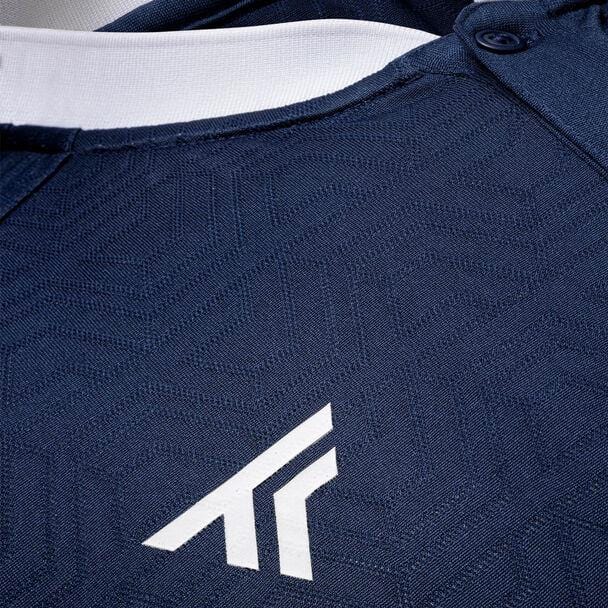 This close-up highlights the dark blue textured fabric of the Tecnifibre Men's Team Badminton Mesh Polo Shirt in Marine, featuring a white geometric logo that resembles an abstract letter 'A'. The polyester mesh of the shirt offers excellent breathability, enhanced by a white buttoned neckline.