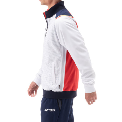 The individual is dressed in a Yonex 70090 Tracksuit Warm-Up Jacket in white with geometric patterns and uses Heat Capsule technology, accented with red and dark blue. They have paired it with matching dark blue Yonex pants, and are photographed in profile against a plain background.