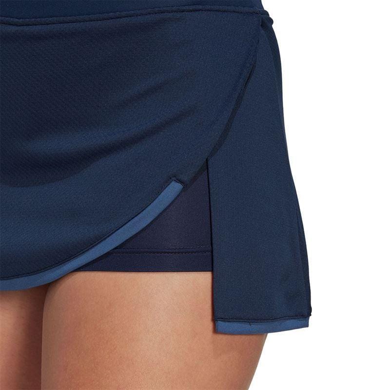 A close-up showcases an individual wearing the adidas Women's Club Badminton Skirt in navy, equipped with AEROREADY technology. This skirt includes built-in short tights and is designed with a curved hem and side slit, elegantly revealing its layered construction.