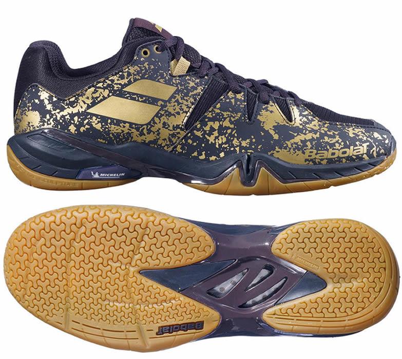 These Babolat Shadow Spirit LTD Men's Badminton Shoes showcase a bold Goldmine Black and Gold design. Equipped with a durable Michelin rubber sole featuring intricate patterns for improved grip, the brand logo is prominently displayed on both the side and sole, providing a perfect blend of style and performance on the court.