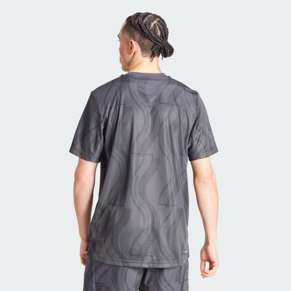 A person with braided hair is wearing an ADIDAS Mens Club Graphic Badminton T-Shirt in Carbon Black, made from recycled materials, along with matching shorts. The outfit incorporates AEROREADY technology for enhanced comfort and is showcased against a light gray background.