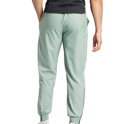 A person is shown from the back wearing green jogger pants and a dark shirt. The ADIDAS Melbourne Men's Pro Badminton Pants by adidas have an elastic waistband and cuffs, and their HEAT.RDY technology ensures the wearer remains cool in white sneakers.