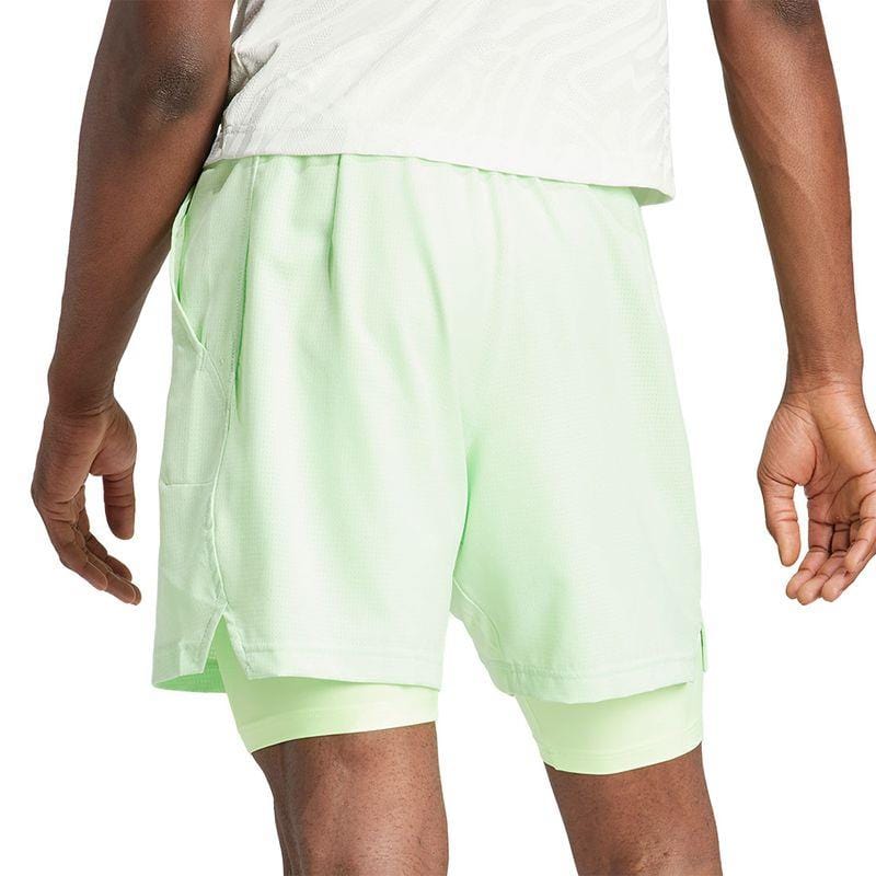 A person is dressed in the green ADIDAS Melbourne Men's 2in1 Badminton Shorts from adidas, paired with a matching shirt. Made using recycled materials, these shorts incorporate HEAT.RDY technology. While outdoors, they subtly bend their left arm, revealing the inner layer visible at the hem of the shorts.