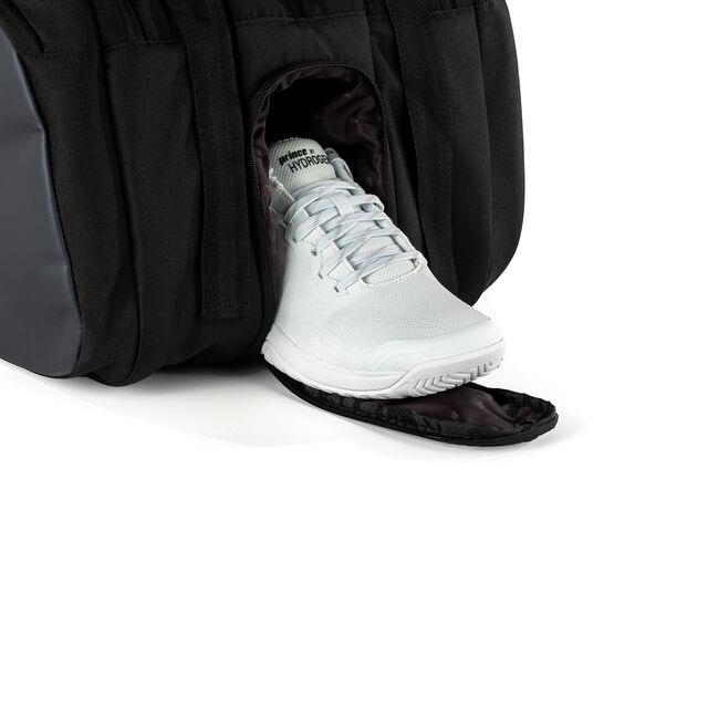 A white sneaker partially emerges from the Prince Tour 3 Comp 12 Racket Badminton Bag by Prince, resting on a dark mat. The bag and sneaker are the primary focus against a plain white background.