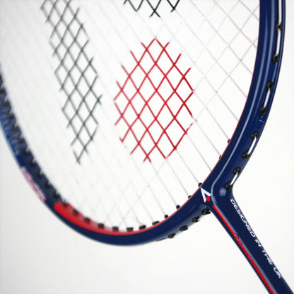 Close-up of the Karakal CB-7 2.1 Badminton Racket, showcasing its blue and red graphite frame. The strings form an intricate pattern, with occasional red strings interspersed among the white ones. The base of the isometric head prominently displays text, highlighting its UK design by Karakal.