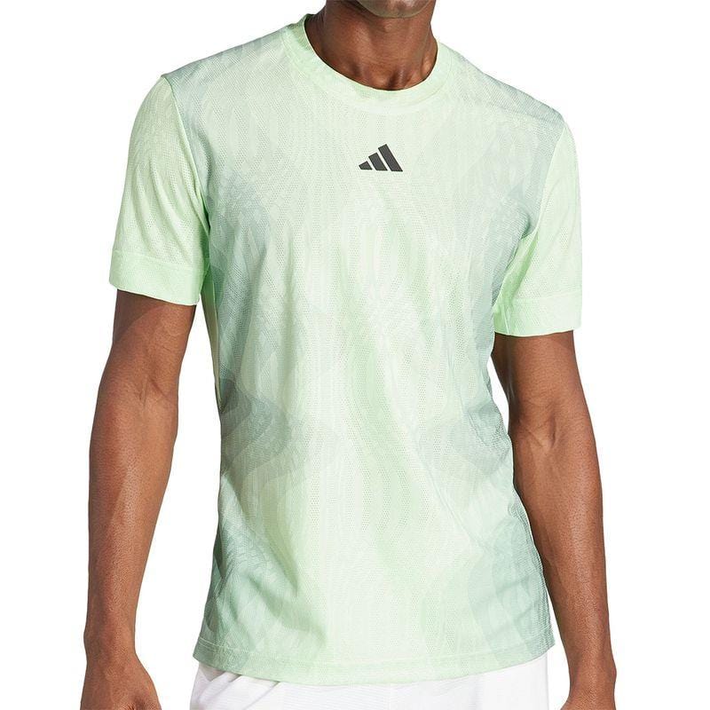 A person is dressed in an ADIDAS Melbourne Men's Pro Badminton T-Shirt in green, showcasing a small black logo and equipped with HEAT.RDY technology for enhanced comfort. Crafted from recycled polyester, the shirt features a discreet pattern and is paired with white shorts. The individual is not entirely visible.