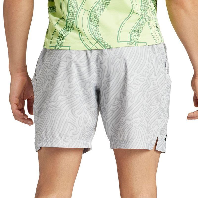 A person is wearing a light green patterned shirt and the ADIDAS Melbourne Men's Pro 7 Inch Badminton Shorts in gray, crafted from recycled polyester featuring a wavy line design. The image focuses on the back view from mid-thigh to waist, emphasizing the comfort of HEAT.RDY fabric that enhances breathability.