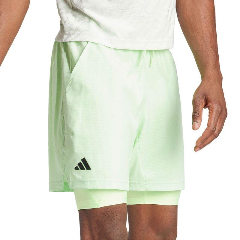 A person dressed in ADIDAS Melbourne Men's 2in1 Badminton Shorts in green, designed with recycled materials. These shorts have a relaxed fit and showcase the iconic logo on the left, layered over compression shorts. The look is paired with a pristine white shirt.