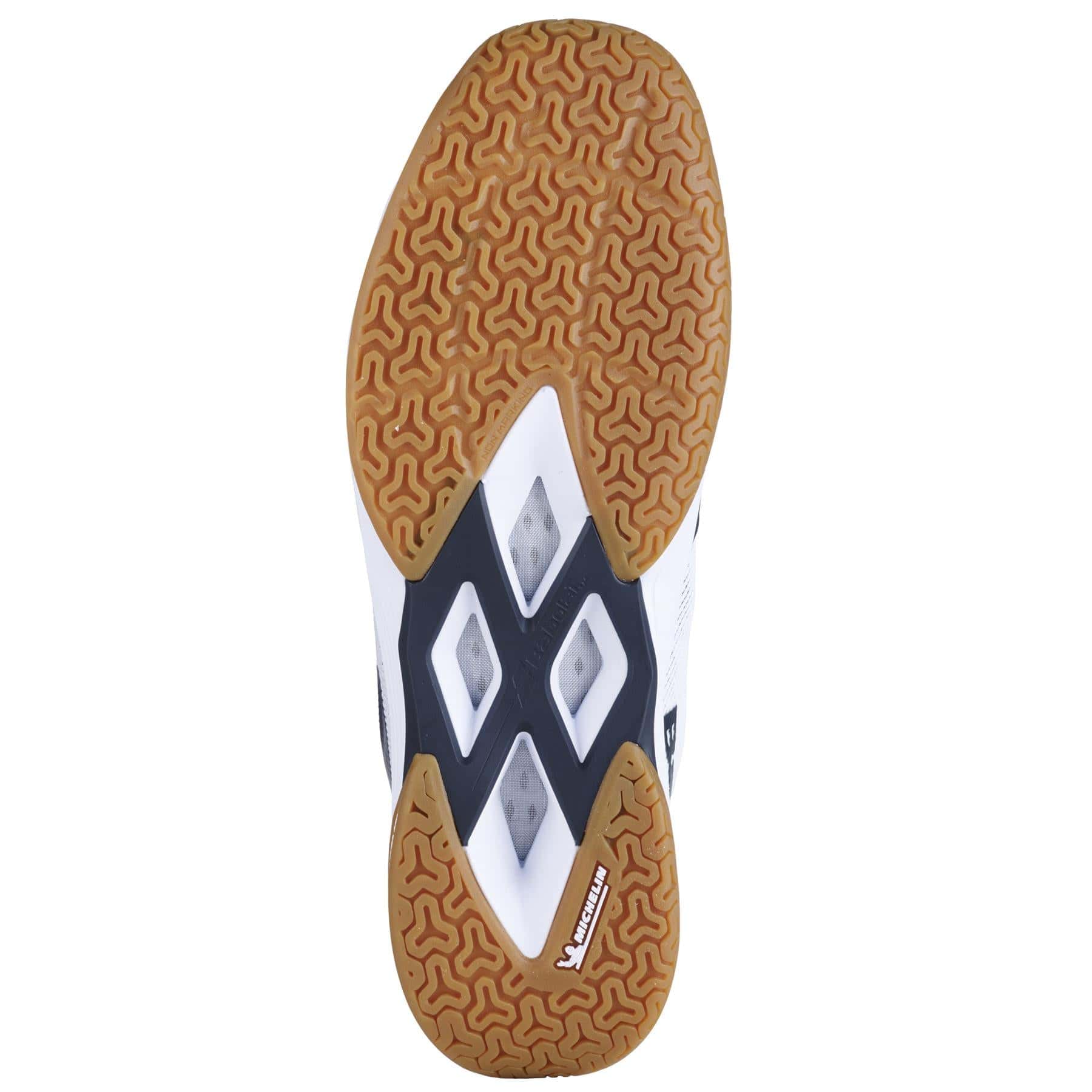 The image showcases the sole of a Babolat sports shoe, designed with Michelin Rubber for improved traction. It features a textured, wavy brown pattern combined with black and white geometric designs and has triangular cutouts in the center. The brand name "Babolat" is printed near the heel.