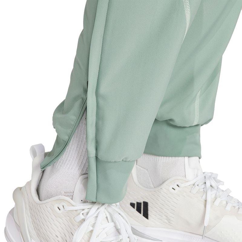 A person wearing adidas Melbourne Men's Pro Badminton Pants in green, equipped with AEROREADY technology, paired with white sneakers.