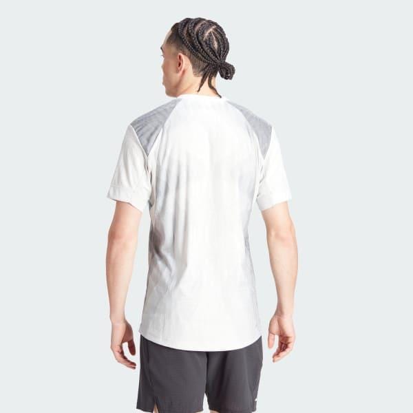 A person with braided hair stands with their back to the camera, dressed in an adidas Melbourne Men's Pro Badminton T-Shirt made with Parley Ocean Plastic and dark, short athletic shorts against a plain, pale gray background.