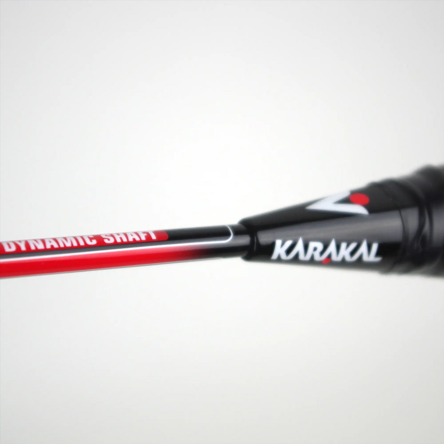 Close-up of the Karakal CB-2 2.1 Junior Badminton Racket handle and shaft, displaying the "Dynamic Shaft" label in red and white. The name "Karakal" appears near the grip, while featuring an aluminum head against a plain, light-colored background.