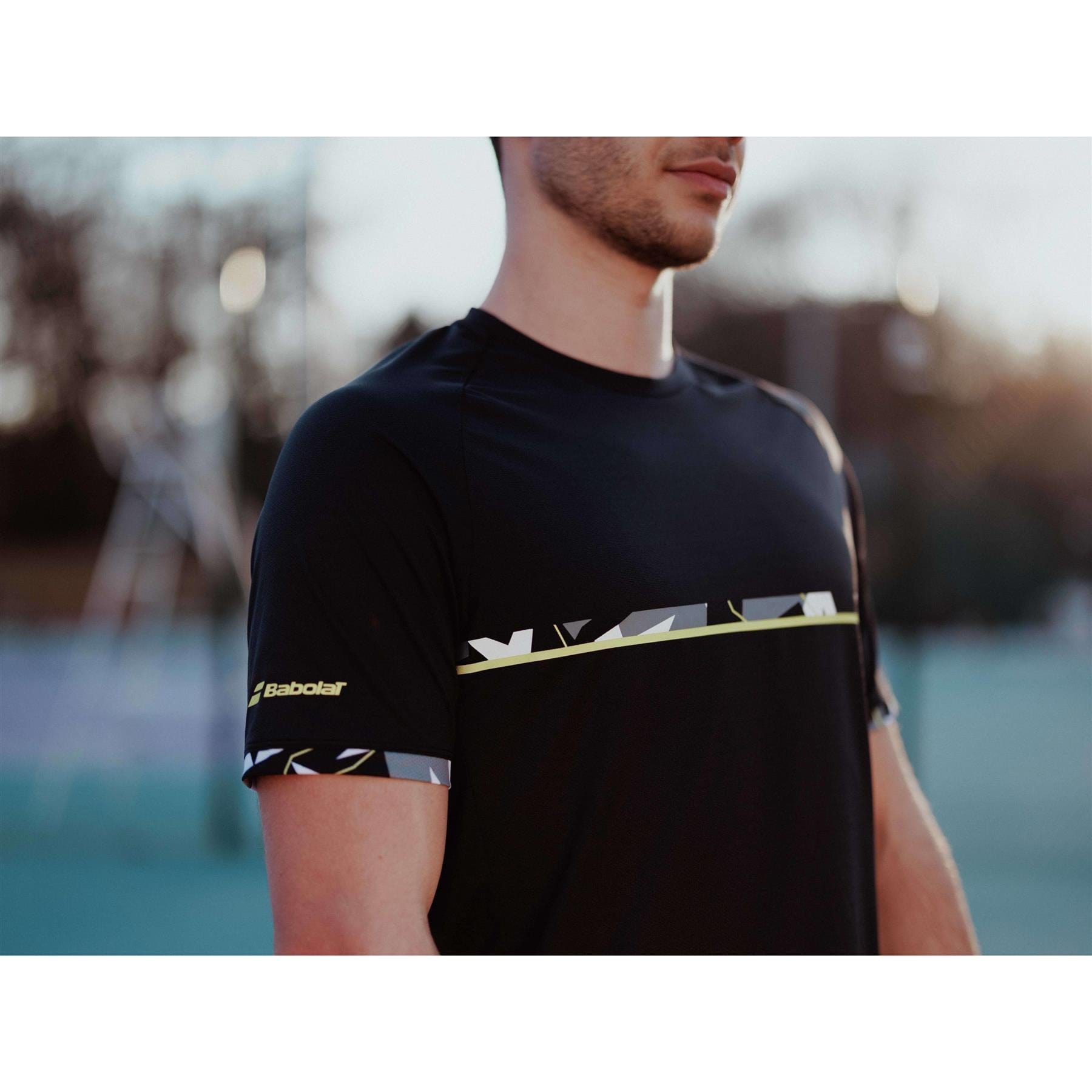 A person wearing a Babolat Aero Crew Neck Men's Badminton T-Shirt in black, adorned with geometric patterns and a logo on the sleeve, stands outdoors. The fashionable Babolat t-shirt, made from Fiber Dry-polyester material, enhances the softly blurred backdrop of a tennis court and surrounding trees.