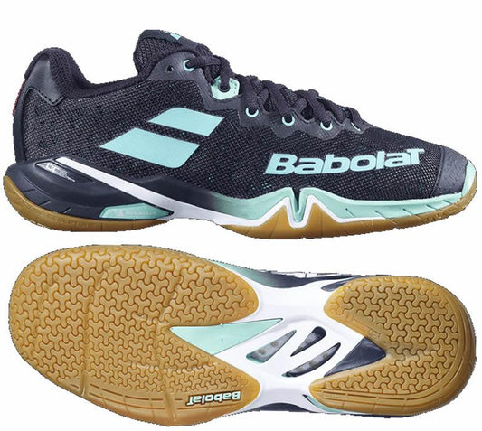 A pair of black and green Babolat Shadow Tour Badminton Shoes, featuring DCS technology and a durable gum sole. The side view prominently displays the brand name, while the bottom view reveals a grip pattern designed for dynamic response and traction on the court.