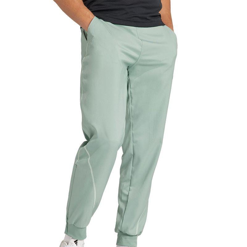 A person is wearing green jogger pants similar to the ADIDAS Melbourne Mens Pro Badminton Pants by adidas, with their hands in the pockets. These pants are highlighted by white piping detail on the sides and are complemented by a dark top and white shoes. The image emphasizes the lower body.
