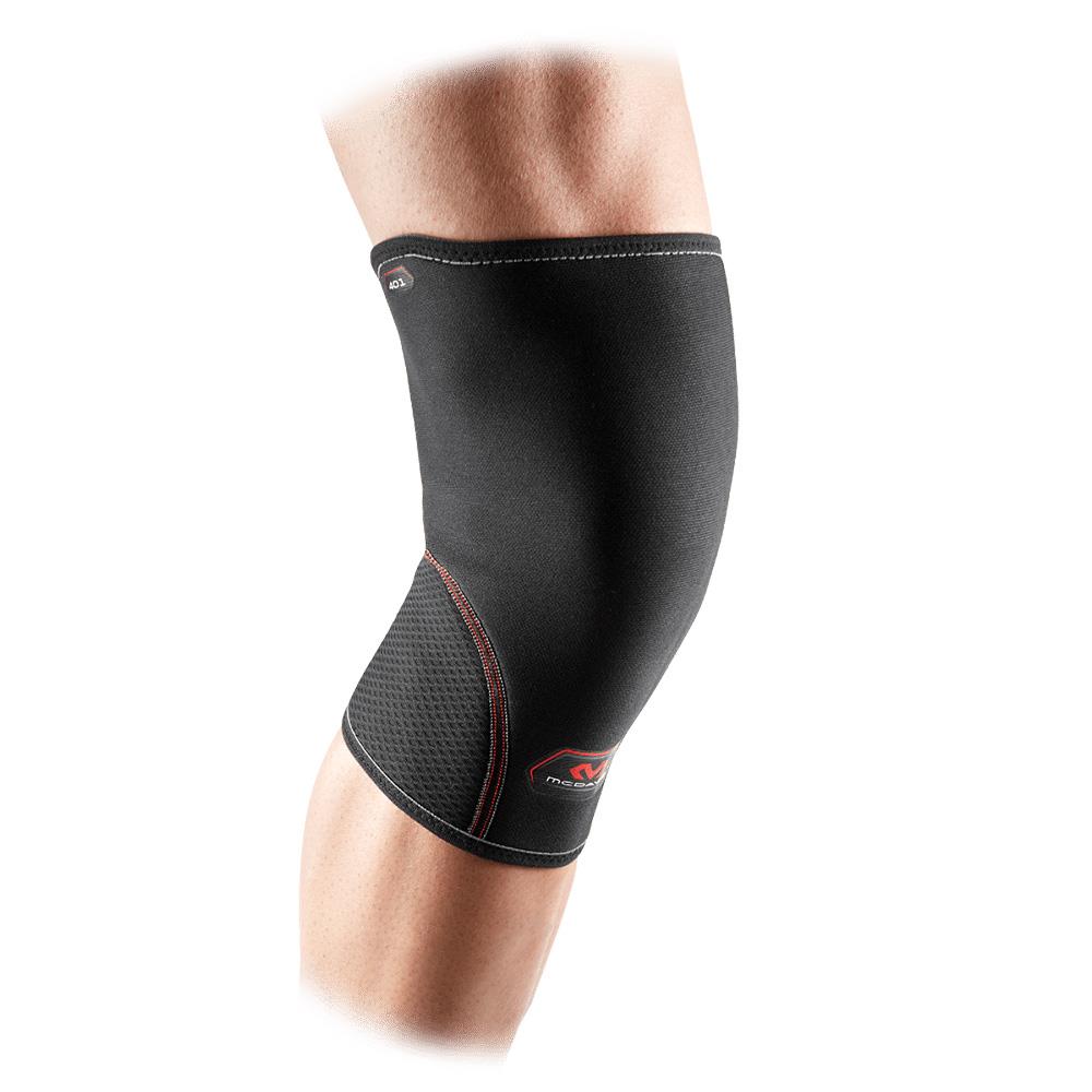 A person wearing a McDavid Neoprene Knee Sleeve, predominantly black with red stitching, tailored for pain relief and compression therapy. The brace presents as smooth and flexible, offering a snug fit around the knee. The background is white.