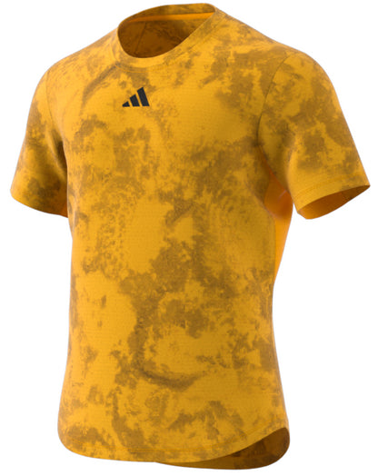 The ADIDAS Paris Men's Freelift Badminton T-Shirt in Gold/Yellow is a short-sleeved piece featuring a textured, marbled pattern. It includes a small black logo on the chest, just below the neckline. The shirt utilizes HEAT.RDY technology for a smooth fabric and comfortable fit, embodying a casual design.