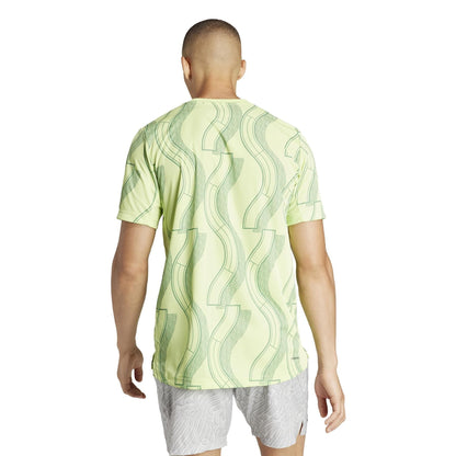 A person is standing with their back to the camera, wearing an ADIDAS Men's Club Graphic Badminton T-Shirt in green from adidas, featuring recycled materials. They are paired with light gray shorts against a simple white background that complements the shirt's AEROREADY comfort.