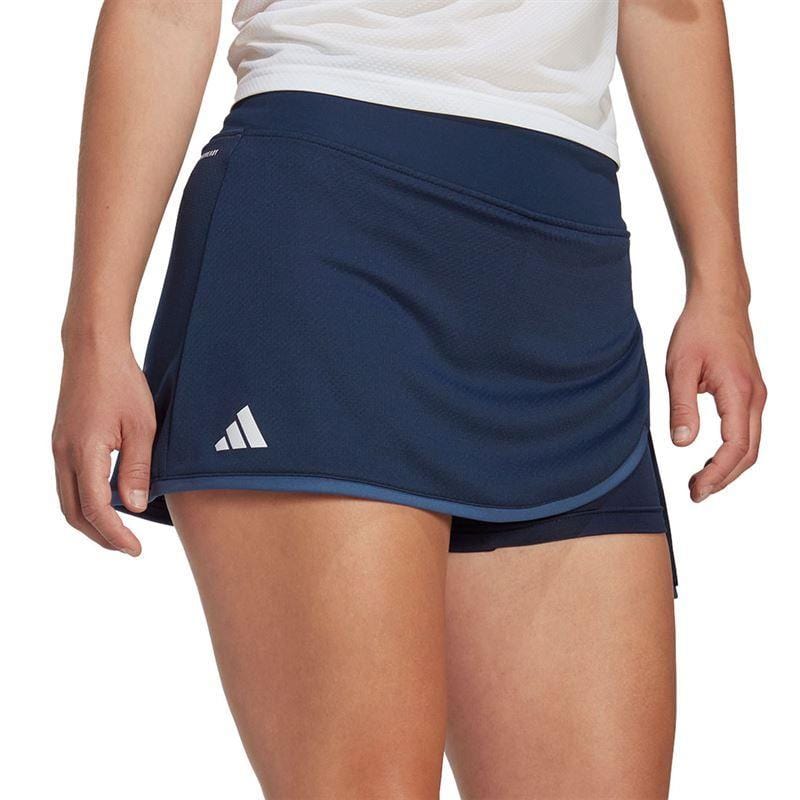 A person is dressed in a navy ADIDAS Women's Club Badminton Skirt, showcasing a small white logo on the left side. This skirt, incorporating AEROREADY technology and built-in short tights, features a curved hem and is complemented by a white top.