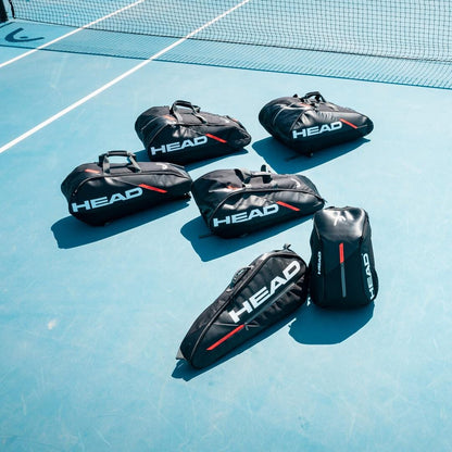 Six black HEAD Tour Team 3R badminton bags are neatly arranged on a blue tennis court. Each bag prominently displays the HEAD brand logo in white with red accents and features an adjustable shoulder strap. A net is visible in the background, completing this perfect setup for serious players.