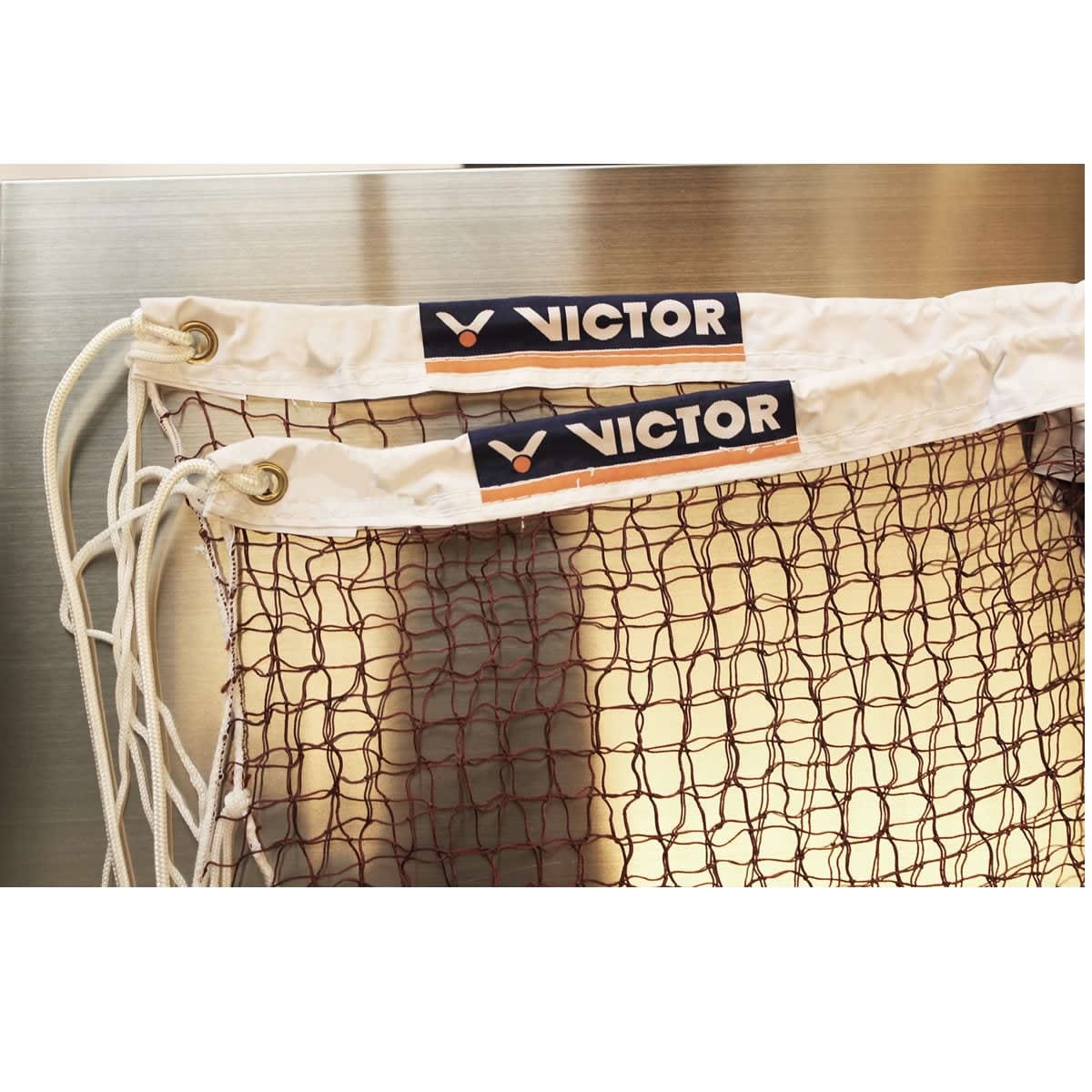 A close-up showcases two Victor Badminton Net International Tournament Level products, featuring white fabric tops tied with nylon cords and boasting the highest quality. The Victor brand logo prominently adorns each net against a smooth, metallic background.