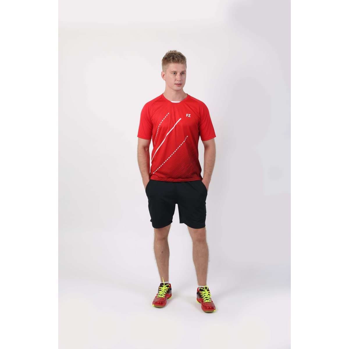 A person stands against a plain background, sporting an FZ Forza Balkan Red Boys/Mens Badminton T-Shirt that offers a comfortable fit. They pair it with black shorts and vibrant red and yellow sports shoes. Their short hair complements the relaxed look as their hands rest at their sides.